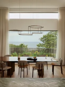 Dining room with a large wooden table and chairs, set with various items, surrounded by a large window with natural light