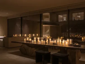 A modern dining room with a candlelit table, chairs, and a large window allowing natural light to enter