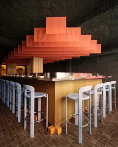 A modern bar with a wooden counter and blue stools, dim lighting, and various items on the counter