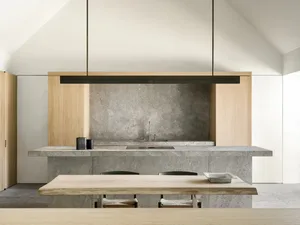 A modern kitchen with a wooden table and marble top, surrounded by black chairs, a sink, and a hanging light, with natural lighting creating a warm atmosphere