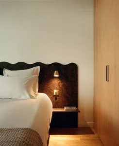 Cozy bedroom with a brown nightstand, a white bed with a brown headboard, and a dimly lit space with a white wall and brown floor