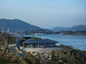 A serene modern house with a stone exterior, metal roof, and large windows, sitting on a hillside overlooking a body of water
