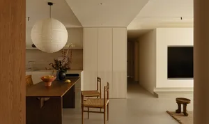 A dimly lit kitchen and dining area with a wooden counter, sink, dining table, chairs, potted plant, vase, and clock, creating a warm and inviting atmosphere