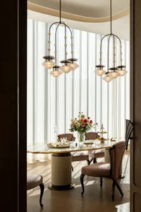 Dining room with a glass table, wood chairs, and a couch in the background, set with a vase of flowers, wine glasses, and a bowl, under soft and warm lighting