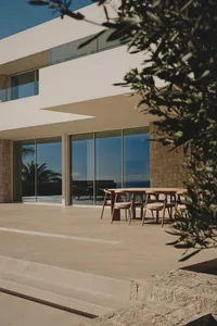 A modern house with a patio area featuring a glass door, large window, and comfortable outdoor seating with a dining table and chairs, surrounded by natural light and visually appealing materials