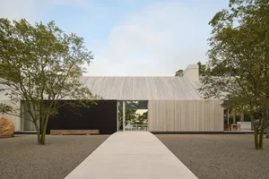 A modern house with a wooden exterior, a large deck, and a covered area, set against a natural outdoor backdrop with sunlight and a black door