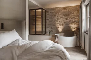 Cozy bedroom with a stone wall, a bed with a white comforter, a bathtub, and a vase with flowers on the window sill