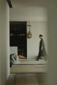 A woman in a black coat walks down a calm hallway lined with cabinets and a stone wall