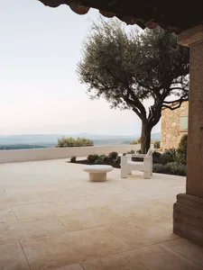 A relaxing outdoor patio with a stone wall, a white bench, and a wooden chair, surrounded by a tree and natural surroundings