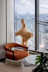 A modern living room with a large window, a wooden sculpture, a glass table, and a potted plant, lit by natural sunlight