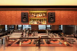 A dimly lit bar area with a wooden counter, stove, and shelf, featuring a variety of bottles and a statue, creating a warm and inviting atmosphere