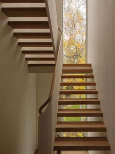 A simple staircase with a white wall and a window on the second floor, providing a view of the outside, with natural light and sunlight coming through