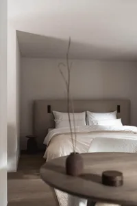 A dimly lit bedroom with a bed, table, and vase, featuring white sheets and a predominantly white color scheme