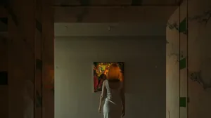 A woman in a white dress admires a painting in a room with a white wall and brown floor