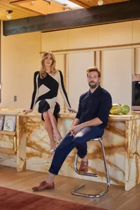 A modern kitchen with a marble counter, dining table, refrigerator, and microwave, featuring a man and woman sitting on the counter
