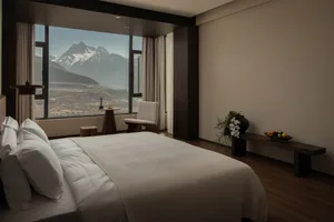 Cozy bedroom with a large window offering a view of mountains, featuring a white comforter, a chair, and a potted plant