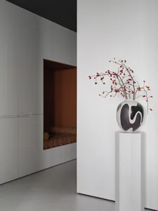 A photograph of a room with a white color scheme, a vase with red flowers on a pedestal, and a doorway in the background