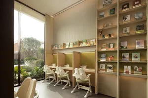 A cozy study area with a wooden floor, white walls, and a large window, featuring comfortable seating and book-filled tables, with soft and warm lighting