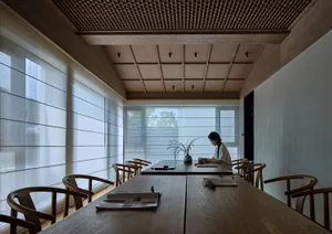 A woman sits at a dining table surrounded by chairs and potted plants in a cozy room with a wooden floor and ceiling beam