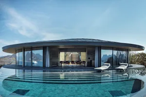 Modern open-concept living space with glass walls, a dining area, two couches, and a TV, surrounded by potted plants and overlooking a pool