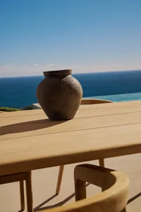 A wooden dining table with a large brown vase on a patio overlooking the ocean, surrounded by chairs in a serene and relaxing environment