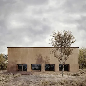 Photograph of a modern building with a brown concrete exterior, a large window, and a bench in a rural field with natural sunlight