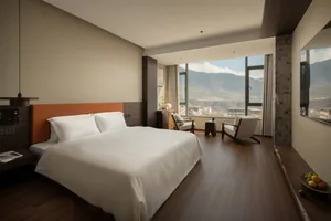 A well-lit bedroom with a wooden floor, a bed, a chair, and a table, near a window