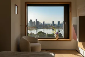 A modern living room with a couch and chair near a large window offering a city view, with a vase and flowers on a table, and natural light creating a bright atmosphere
