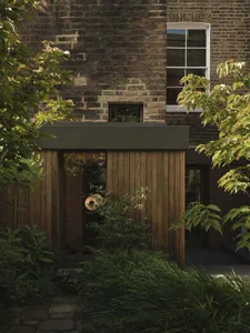 A two-story house with a brick exterior, wooden door, and window, set in a garden with a tree and bush, exuding a calm and serene atmosphere