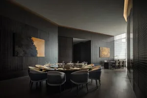 A modern dining room with a large table set with wine glasses and surrounded by chairs, featuring a couch and a potted plant in a dimly lit space with a cozy atmosphere