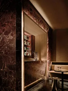 A dimly lit bar area with a marble bar, wooden counter, and bottles on display, set against a background of a sink and a couch in a modern and elegant restaurant
