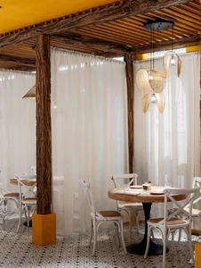 A modern restaurant dining area with white tables and chairs, surrounded by cups, bottles, and bowls, under a mix of natural and artificial lighting