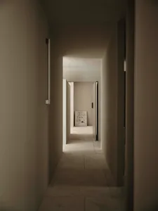 Dimly lit hallway with a white wall, open doorway, and a room beyond, featuring clean lines and simplicity