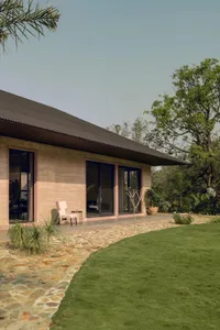 Photograph of a house with a stone exterior, a black roof, a patio with a dining table and chairs, and a garden with a tree and a bench