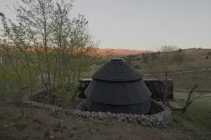 A rustic house with a black roof and a circular structure on top, situated in a wooded area during sunset