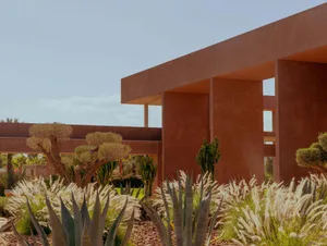 Photograph of a modern building with a red brick exterior, surrounded by lush plants and trees, and a large window allowing natural light to enter