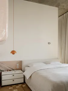 A minimalist bedroom with a white bed, dresser, and lamp, well-lit by natural light through the window