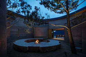 A serene outdoor space with a stone building, a fire pit surrounded by a circular stone wall, and a nearby bench, lit by the warm glow of the fire pit