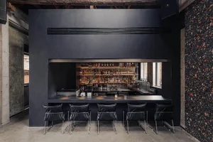 A dark and intimate bar with a long wooden counter and numerous bottles, surrounded by chairs for patrons