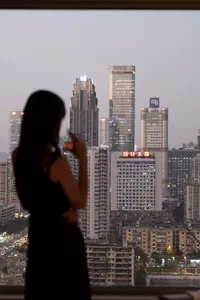 A woman stands on a rooftop at night, admiring the city's vibrant and lively atmosphere, illuminated by artificial lights