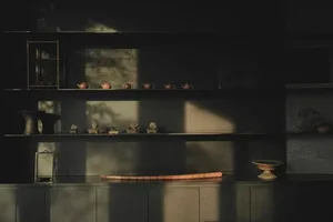 A dimly lit kitchen or dining area with a wooden shelf displaying various objects, a wooden table, and a potted plant, creating a warm and inviting atmosphere