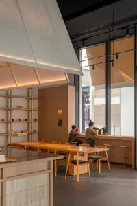 A modern restaurant interior with a dining table, chairs, counter, and kitchen elements, featuring wooden accents and a neutral color palette, with soft and warm lighting