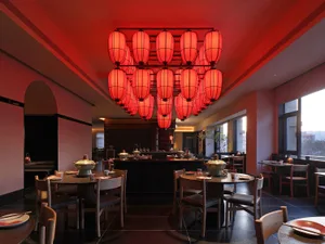 A dimly lit restaurant dining area with a red ceiling, a long table, and various table settings, creating a cozy and intimate atmosphere