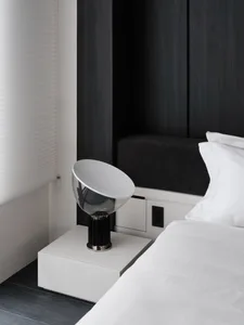 A dimly lit bedroom with a bed on the right, a bench on the left, and a table with a lamp in the middle