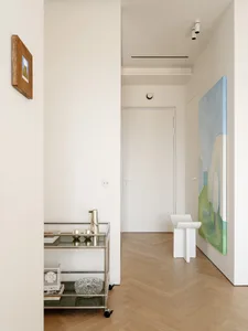 A minimalist hallway with white walls, a wooden floor, a clock on the wall, and a vase on a table, lit by natural light
