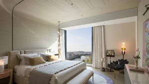 A well-lit bedroom with a large bed, a bench, and a window, featuring a modern style with white and brown colors, wood, and fabric materials
