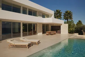 A modern house with a white exterior, a large patio, and a swimming pool surrounded by a deck and palm trees, with natural light and a relaxed atmosphere