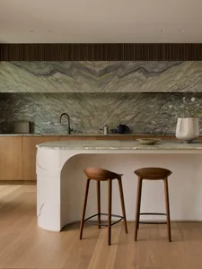 A dimly lit kitchen with a marble countertop, sink, and backsplash, accompanied by wooden stools and a potted plant, creating a cozy atmosphere
