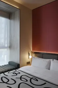 A modern hotel bedroom with a white bed, a bench, and a window, featuring a predominantly white color scheme and natural lighting