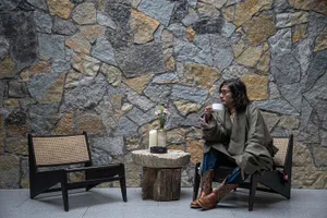 A man sits in a chair in front of a stone wall, holding a cup and surrounded by a vase with a flower on a table